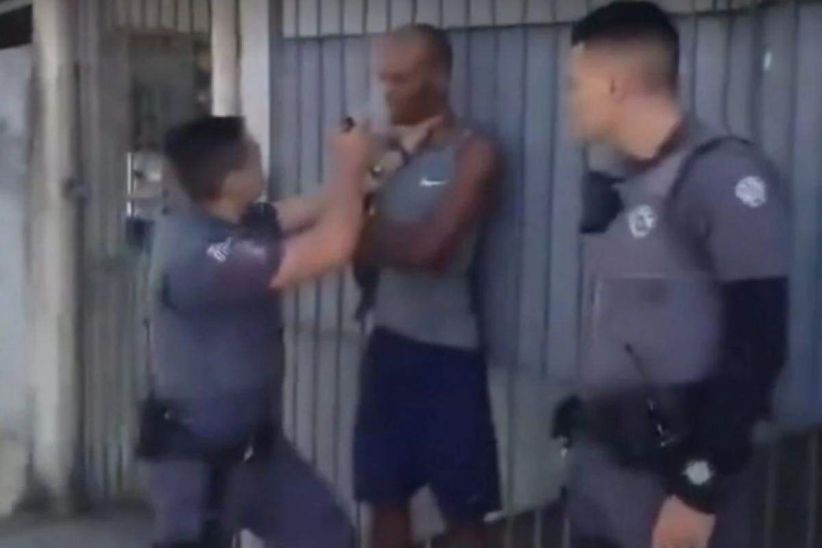 As imagens mostram homem rendido por dois agentes no portão de uma casa; com uma mão no pescoço do abordado, um dos PMs joga spray de pimenta no rosto do homem