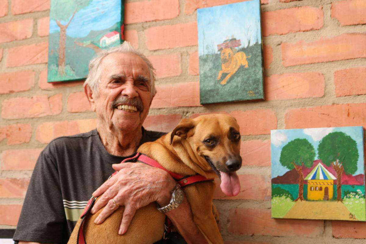 Adairton Ferreira da Costa, de 92 anos, e seu cão Tonico, fonte de inspiração para seus quadros (ao fundo)