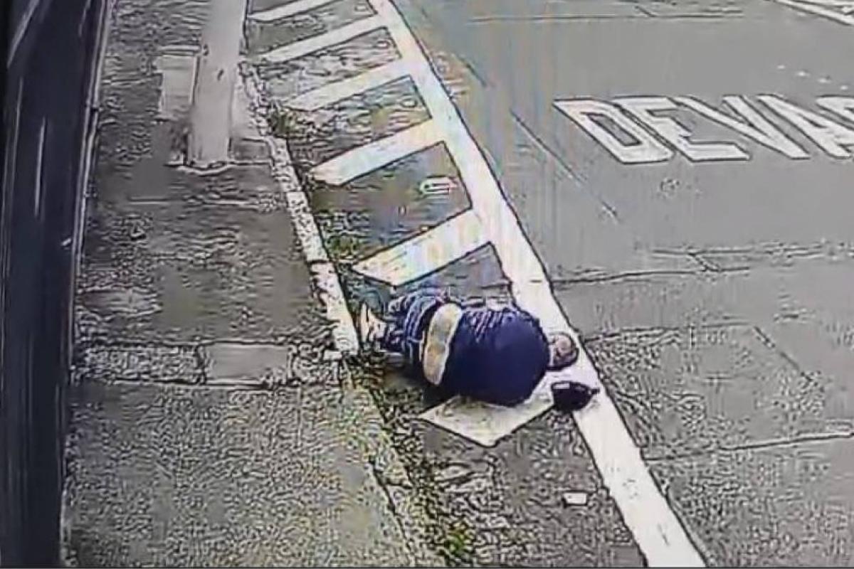 Imagem mostra uma pessoa caída na calçada, vestindo um casaco azul e calças escuras